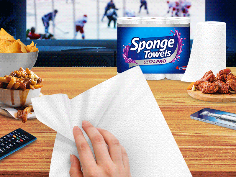 A hand holding a Sponge Towel wipes away a mess on a kitchen counter. There are food items on the counter, a package of Sponge Towels, and a hockey game playing on TV in the background.